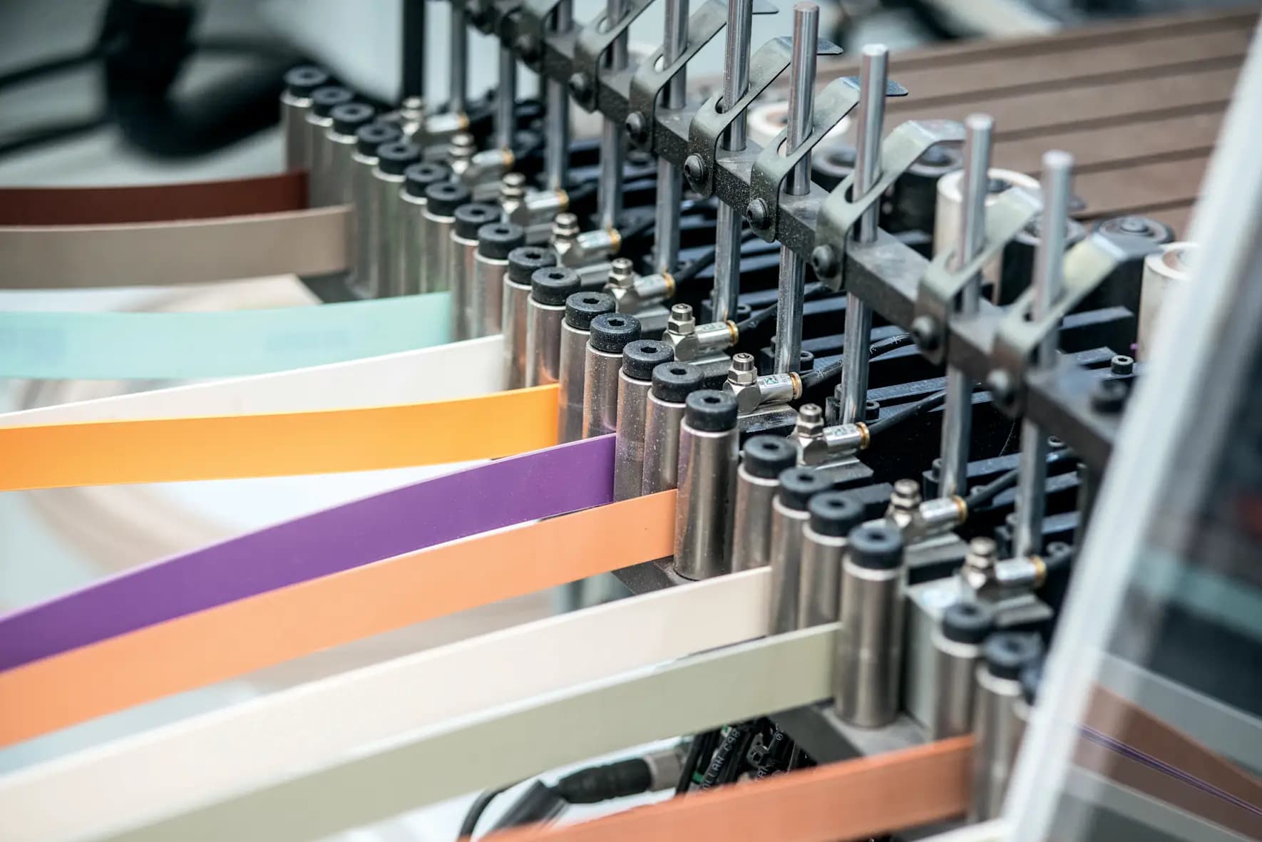 Cintas de canto de colores entrando a una hilera de cilindros neumáticos de una pegadora de canto, con una superficie marrón al fondo.
